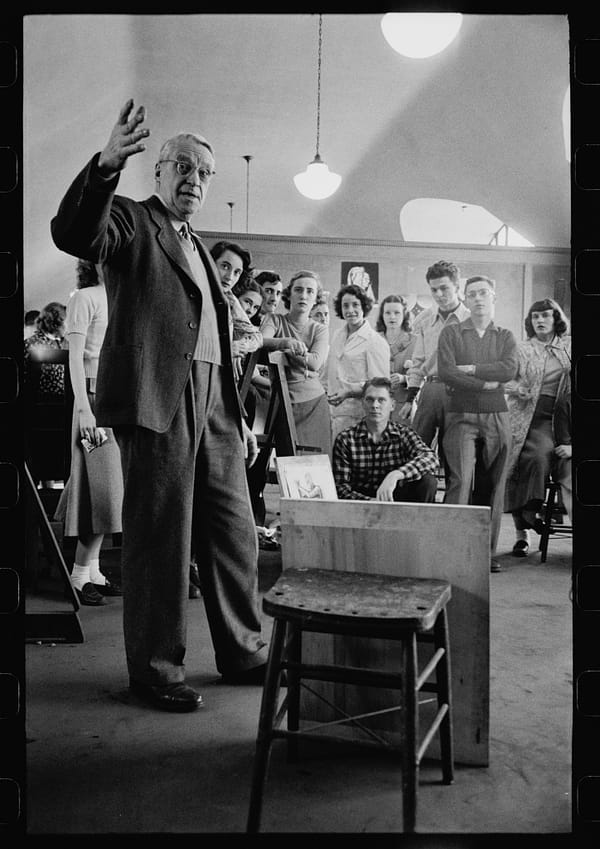 A black and white photo of a professor lecture in an art gallery with students behind him